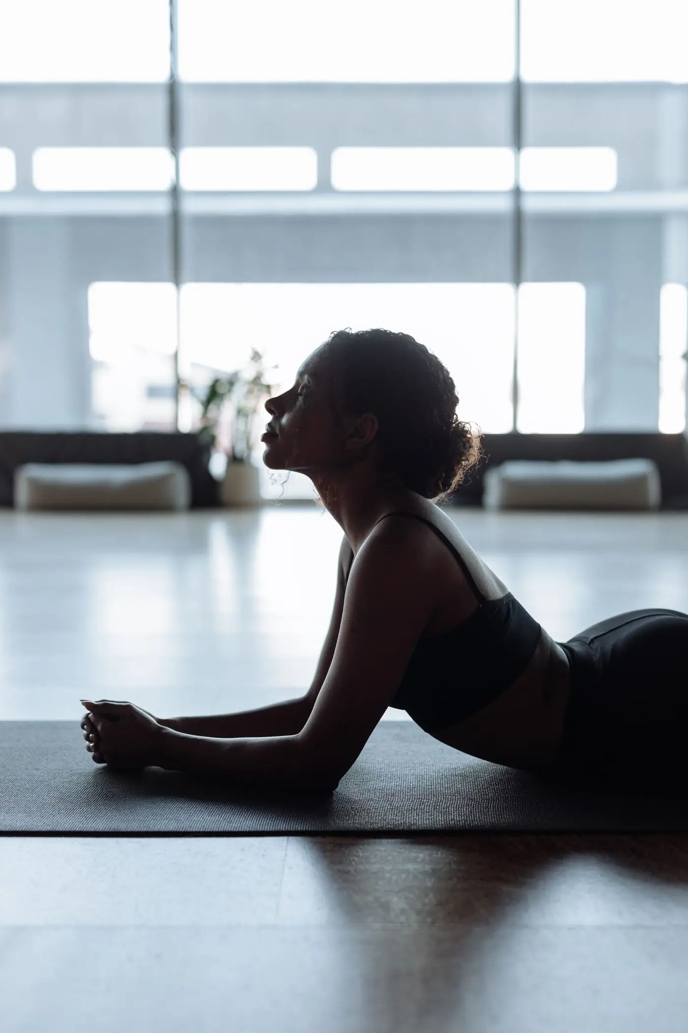 A woman stretching on her mat in a bright, high-windowed studio, backlit